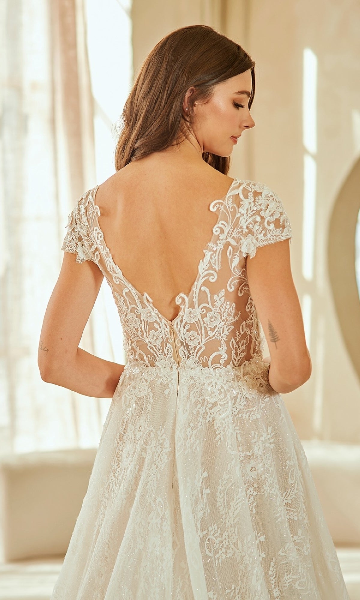 A woman, seen from behind indoors, wears the Amelia Couture 2142 long wedding dress—an A-line gown in white lace with a deep V-back, embroidered cap sleeves, and a corset bodice.