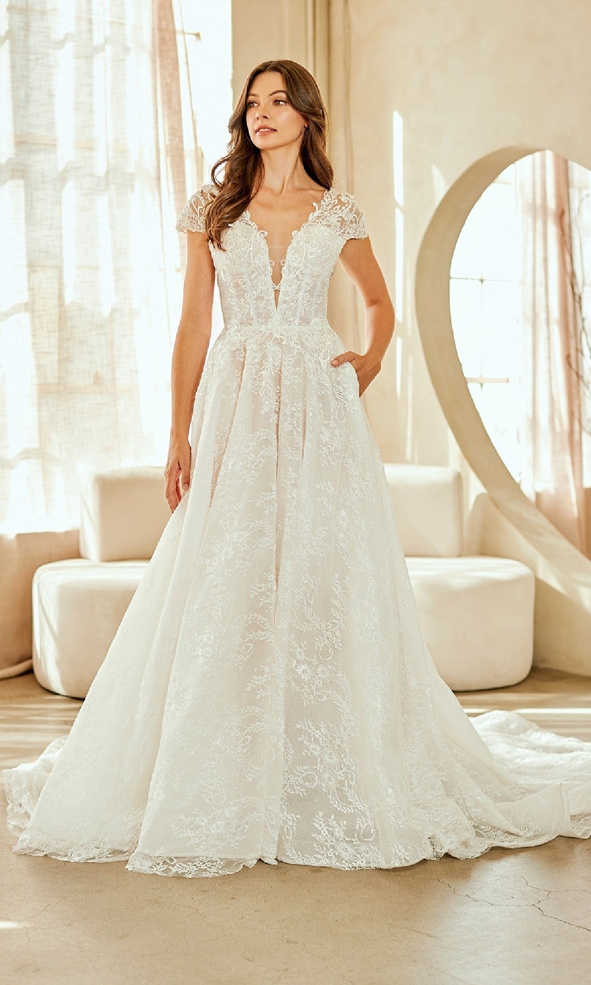 A woman stands indoors in the Amelia Couture 2142 long wedding dress—an A-line gown with lace, cap sleeves, deep V-neck, and a full skirt—while light filters through sheer curtains behind her.