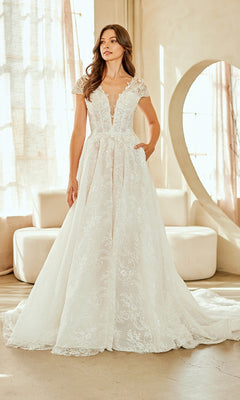A woman stands indoors in the Amelia Couture 2142 long wedding dress—an A-line gown with lace, cap sleeves, deep V-neck, and a full skirt—while light filters through sheer curtains behind her.