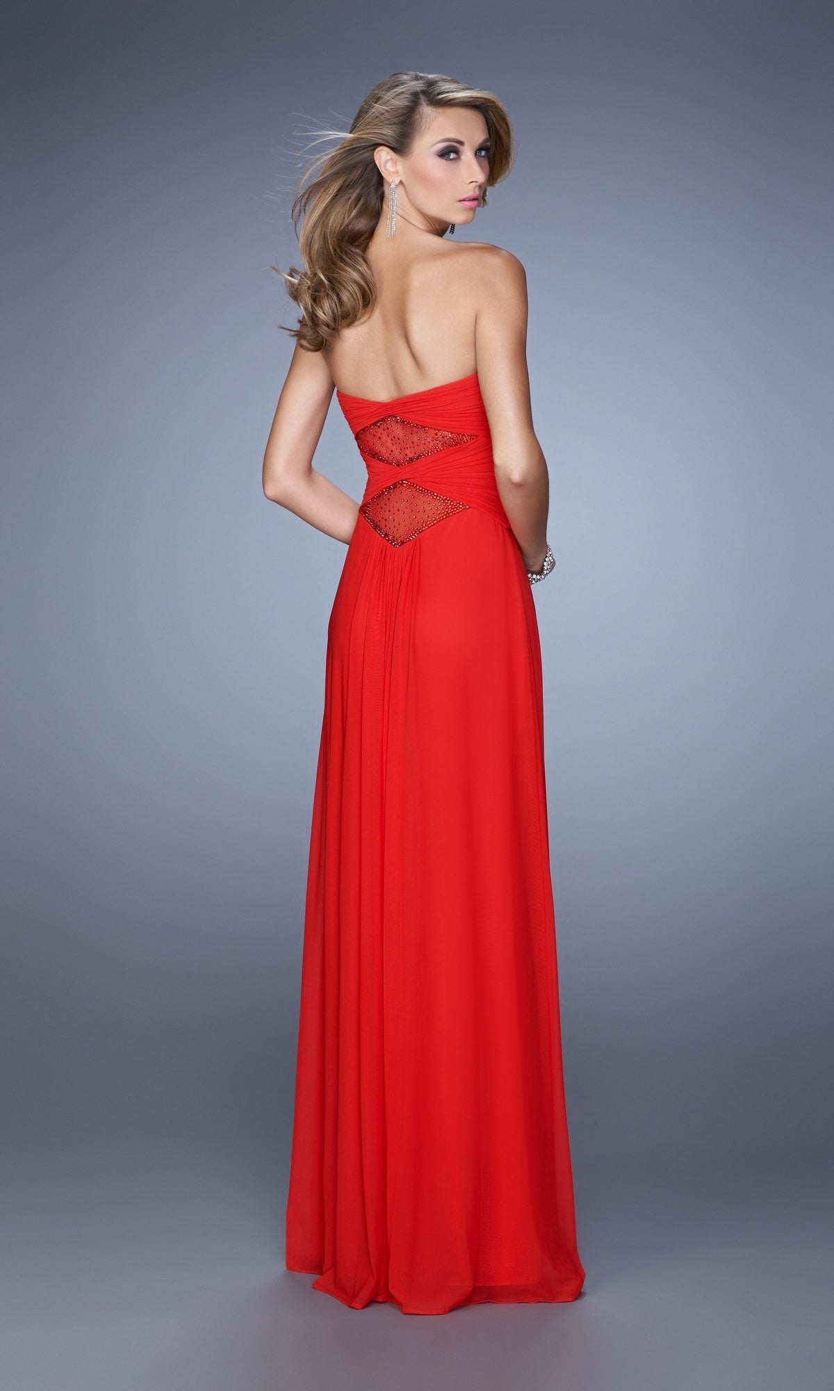 A woman stands facing away against a gray background, wearing the La Femme Dress 21462—a strapless, floor-length red gown featuring a ruched bodice and sheer embellished back cut-outs.