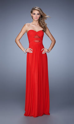 A woman poses with one hand on her hip in the La Femme Dress 21462, a strapless, floor-length gown featuring a ruched bodice and embellished cutouts, set against a gray background.