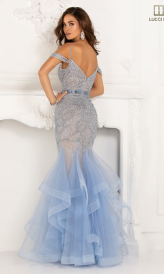 A woman stands before a white ornate fireplace in the Lucci Lu 2147, an open-back mermaid prom dress featuring light blue beaded lace embellishments and a layered tulle skirt.