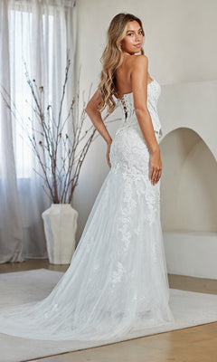 A woman wearing the Amelia Couture 2151 Long Prom Dress, featuring a strapless corset bodice with elegant lace embroidery, a fitted silhouette, and train, stands indoors looking over her shoulder.