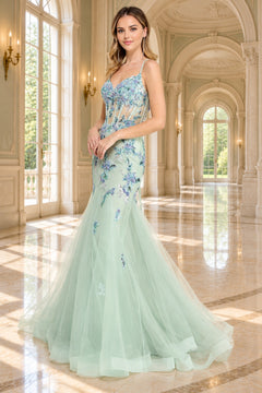 A woman wears the Amelia Couture 2164, a sleeveless, mint green mermaid prom dress with a corset-style bodice featuring blue sequin flower appliques and a tulle skirt.