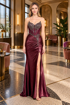 A woman wears a deep burgundy Long Prom Dress: Amelia Couture 2166, a strapless, floor-length gown featuring a beaded corset bodice and a high side slit.