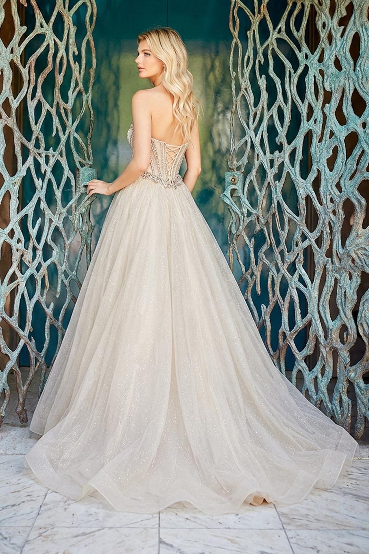 A woman wears a Long Prom Dress: Amelia Couture 2172, a strapless champagne gown with a lace-up bodice and a full, sparkling tulle skirt, standing in front of a decorative metal gate.