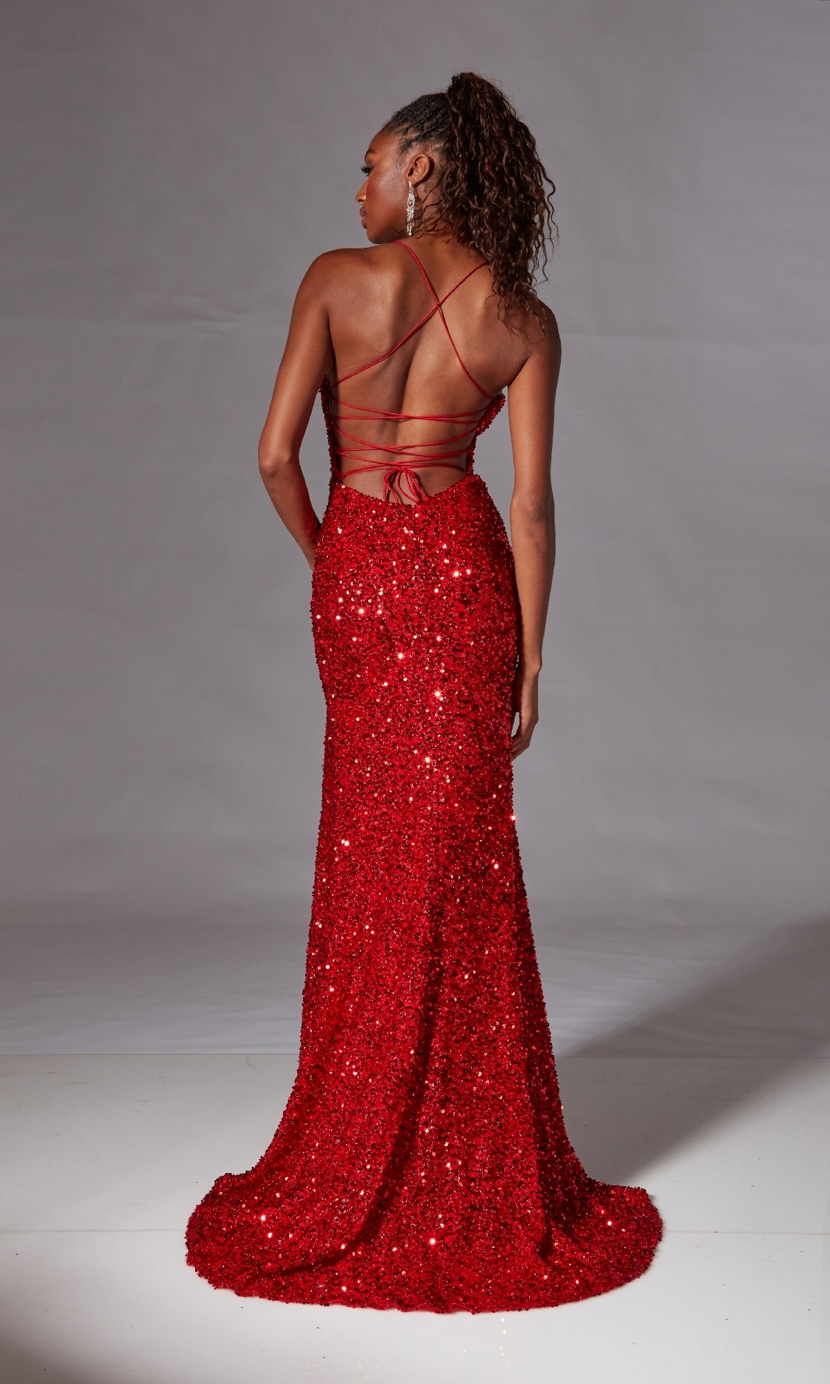A woman stands facing away against a plain backdrop, wearing the Backless Long Sequin Prom Dress: Aerie Couture 217—a floor-length, red gown with an open strappy back and shimmering sequins.