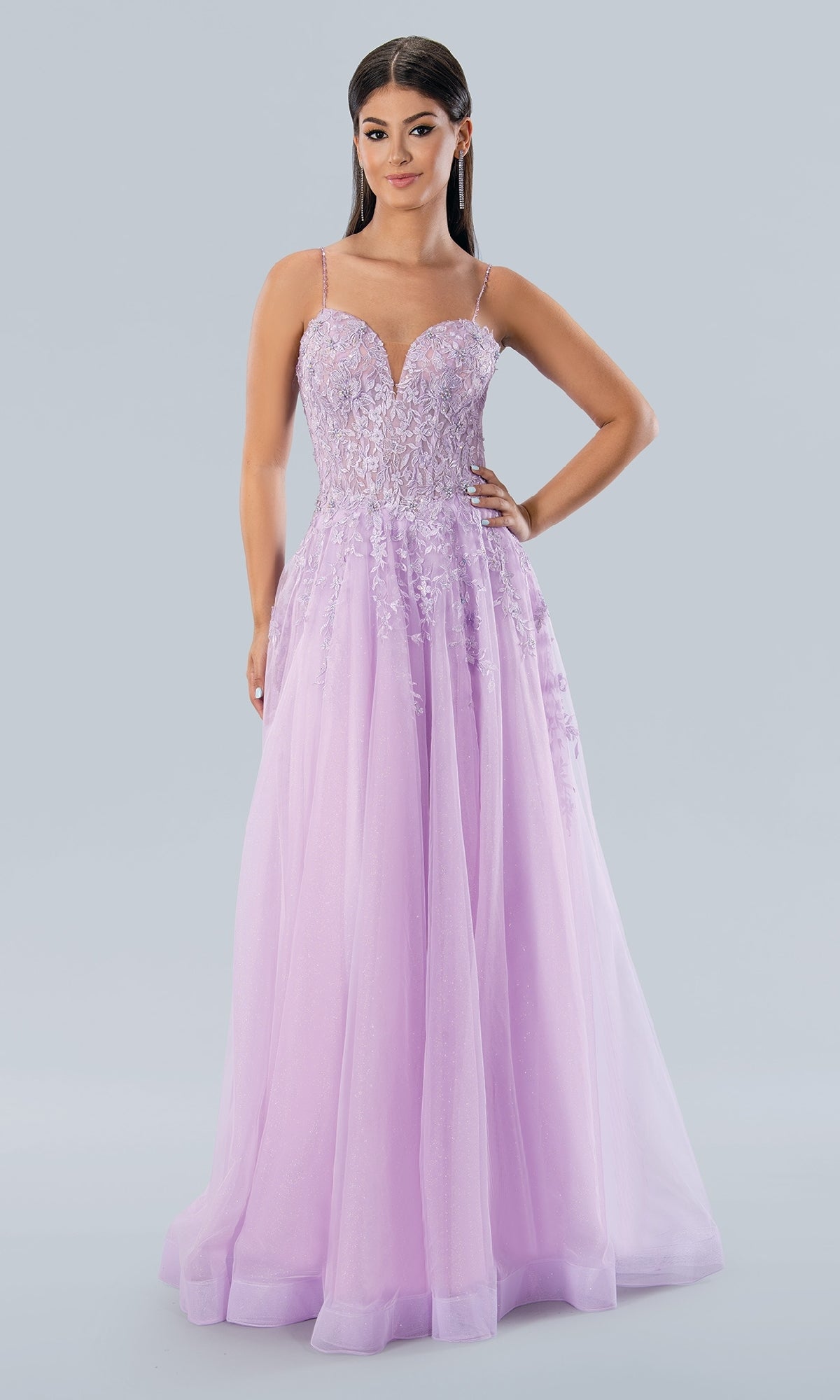 A woman wears the Stella Couture 22027, a long lavender glitter tulle prom dress with thin straps and a beaded fitted bodice, standing against a plain light background.