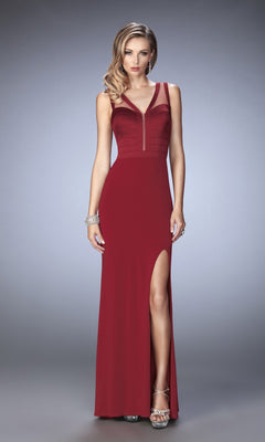 A woman stands against a gray background wearing the La Femme Dress 22301—a sleeveless, floor-length maroon gown featuring a high slit, sheer neckline detail, and satin bandage-style bodice—paired with a silver bracelet and strappy heels.