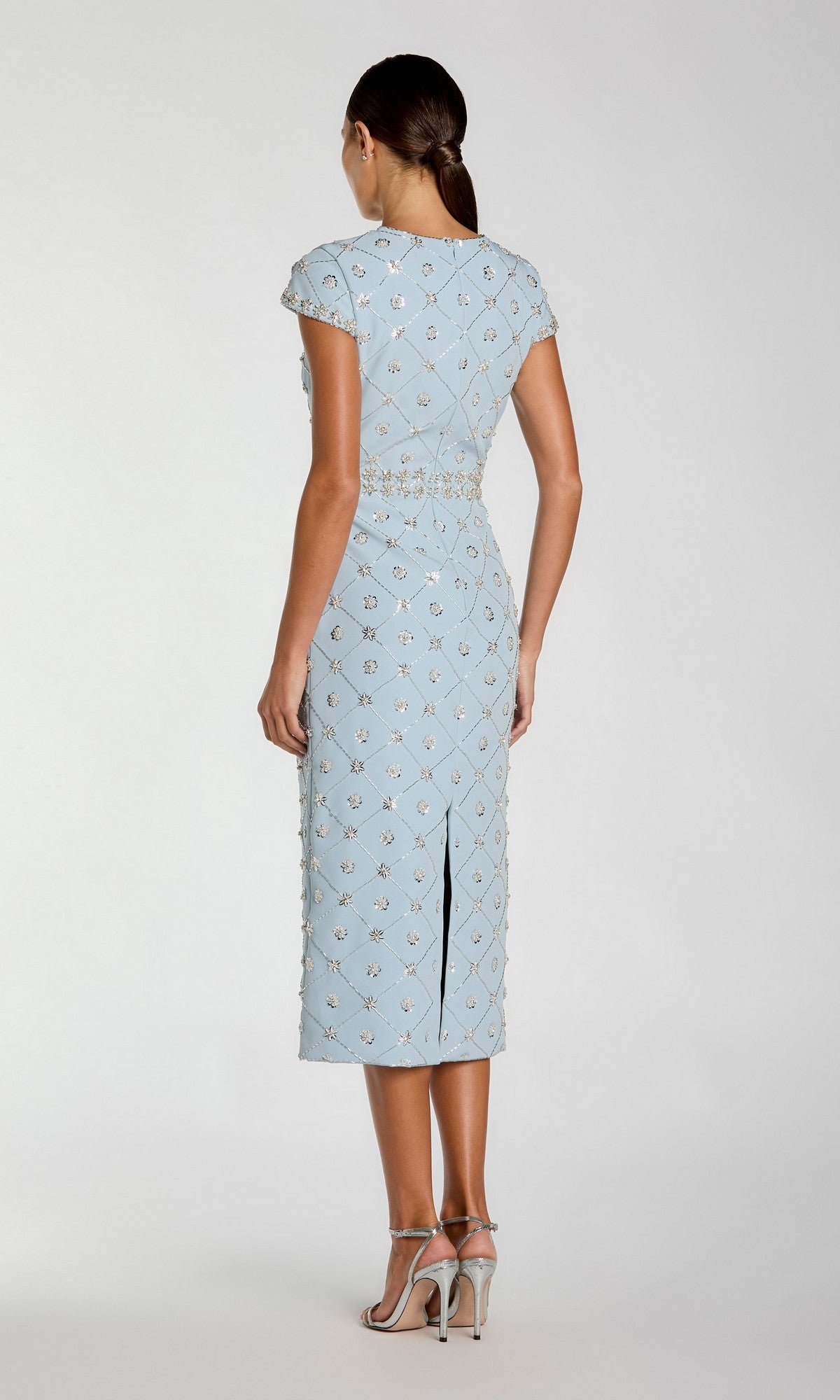 A woman stands facing away, wearing the light blue Midi Beaded Wedding Guest Dress: Mac Duggal 2234 with silver embellishments and high-heeled sandals.