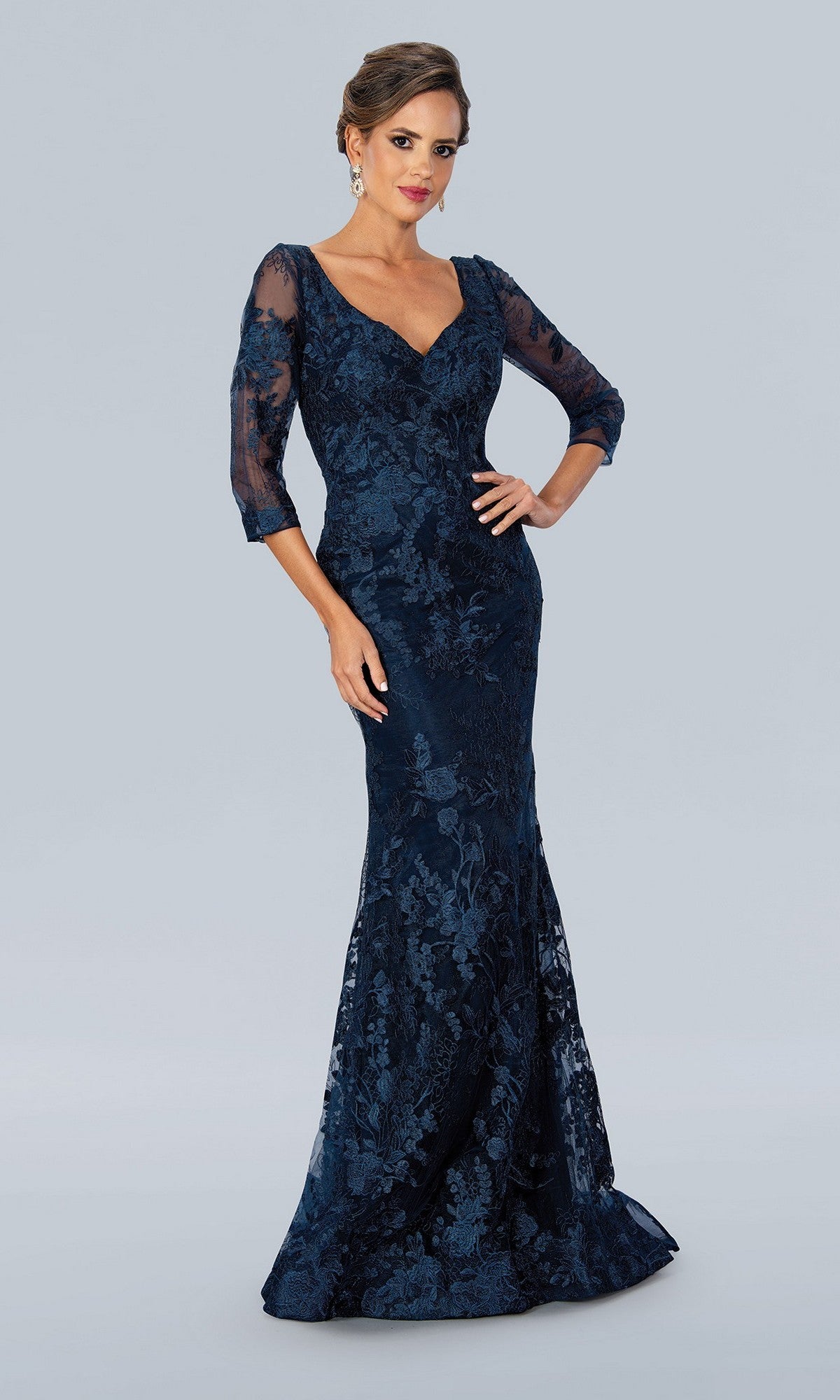 A woman models the Stella Couture 22361 long formal dress in navy, featuring a v-neckline, three-quarter sheer sleeves, and intricate lace embroidery, standing before a plain light gray background.