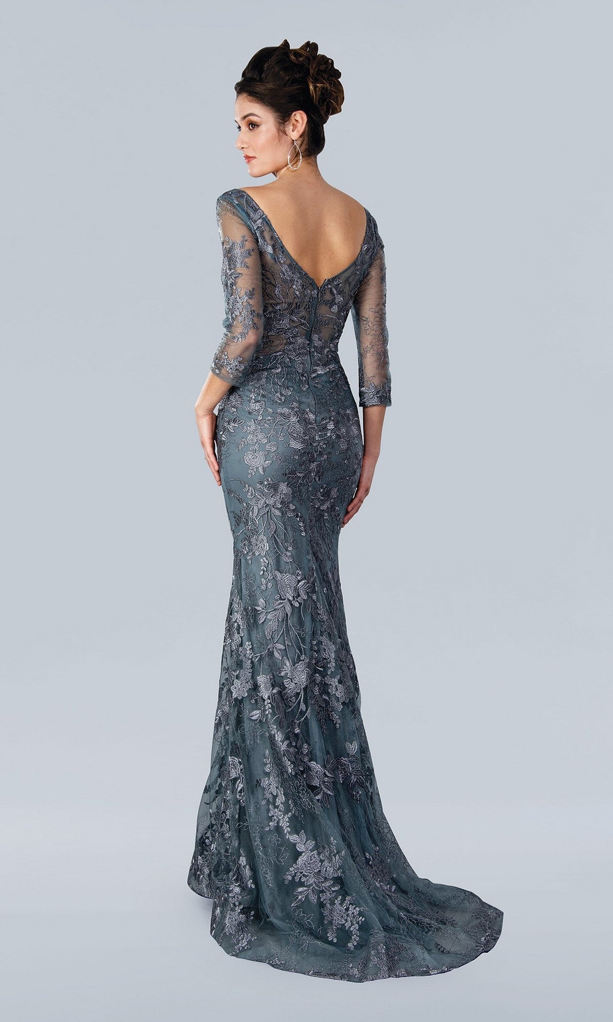 A woman models the Stella Couture 22361 long formal dress in charcoal with a fitted lace design, sheer sleeves, and an open back, standing against a plain light gray background.