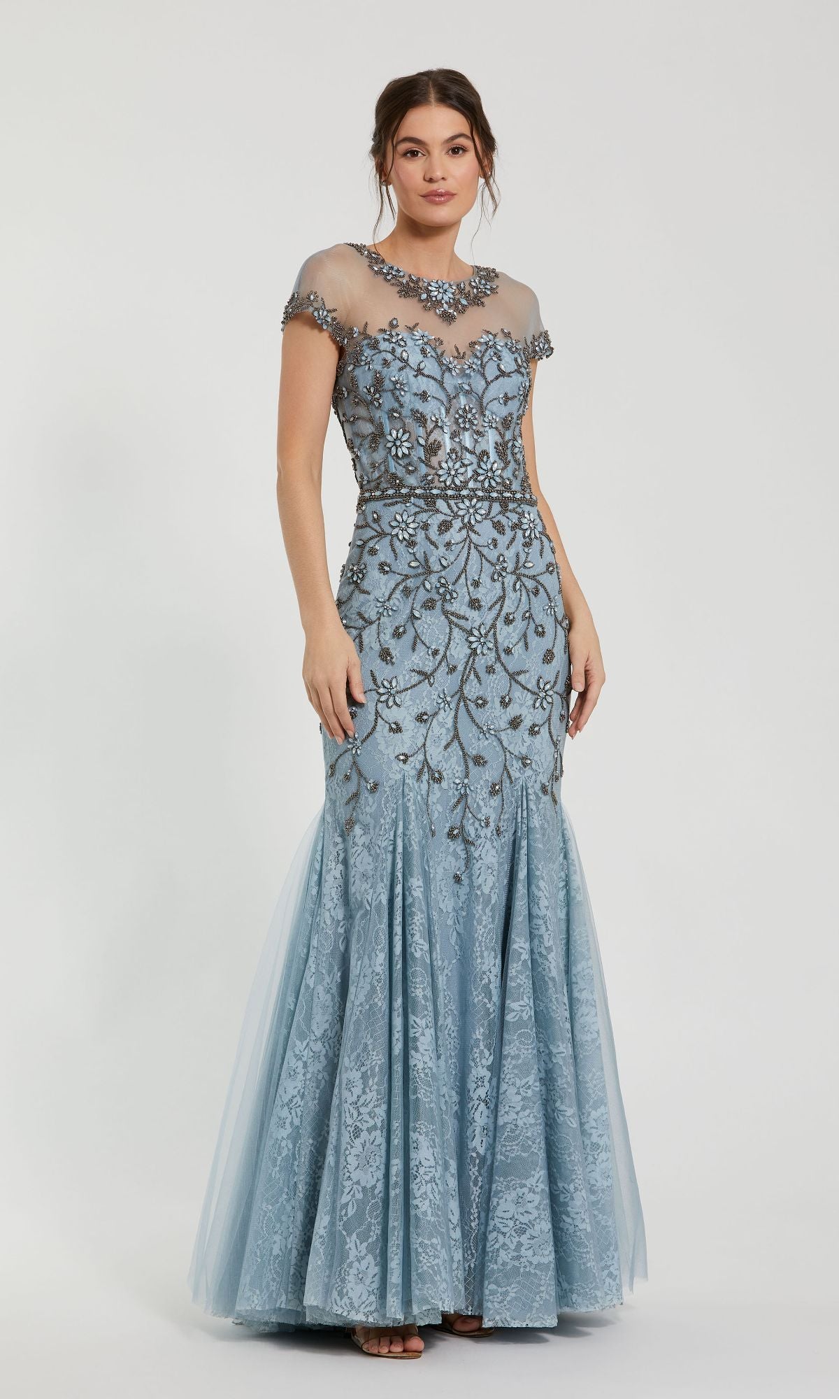 A woman stands gracefully against a white background wearing the Beaded Long Blue Formal Dress: Mac Duggal 2239, featuring cap sleeves, a sheer neckline, and intricate silver embroidery.