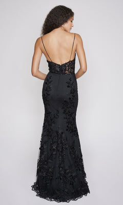 Woman in the Nina Canacci 2240 long formal dress—black, floor-length, mermaid silhouette with a backless design, thin straps, and floral embroidery—stands against a plain light background, facing away from the camera.