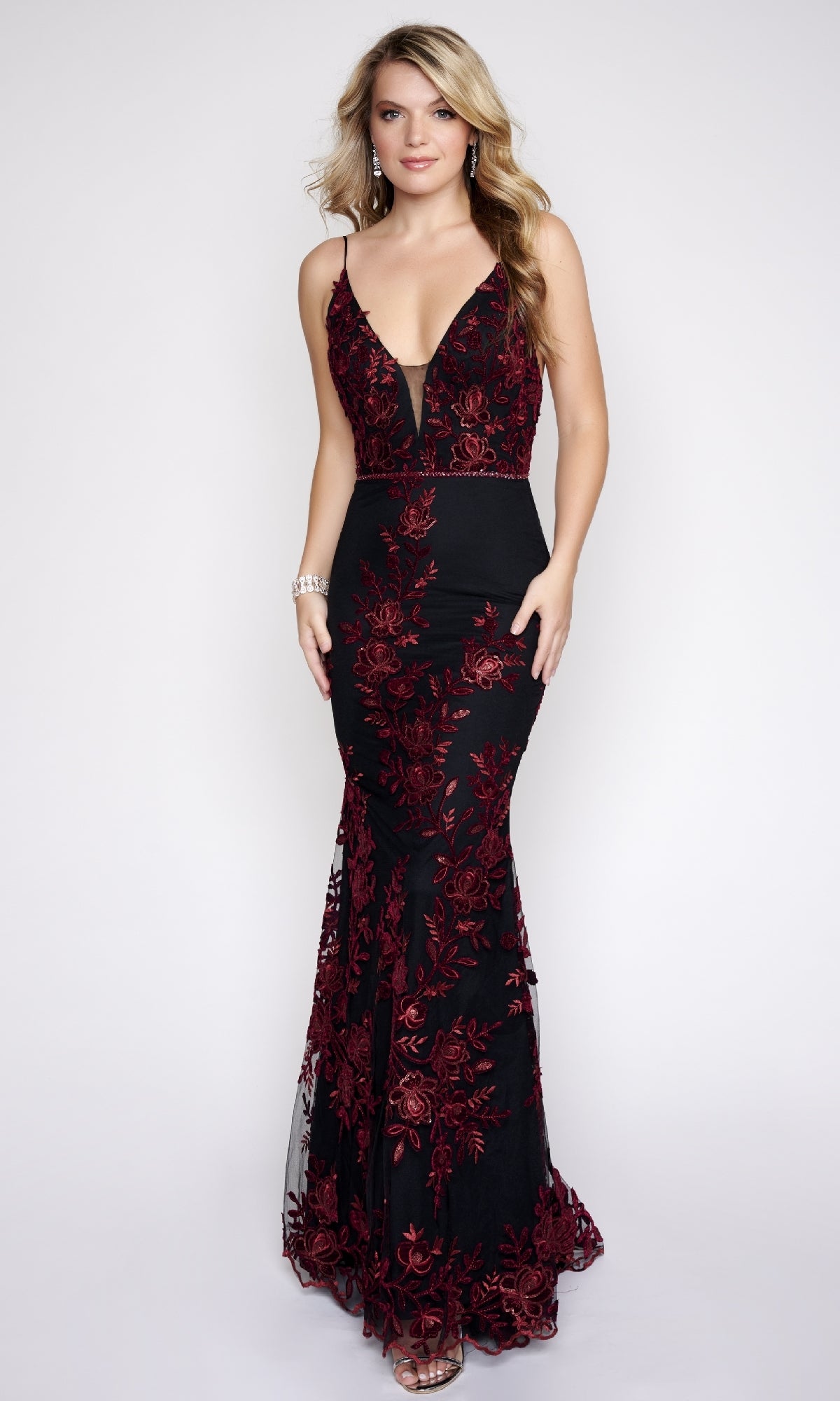 A woman stands against a plain light background wearing the Nina Canacci 2240, a long formal black mermaid gown with red floral embroidery, spaghetti straps, and a deep V neckline.