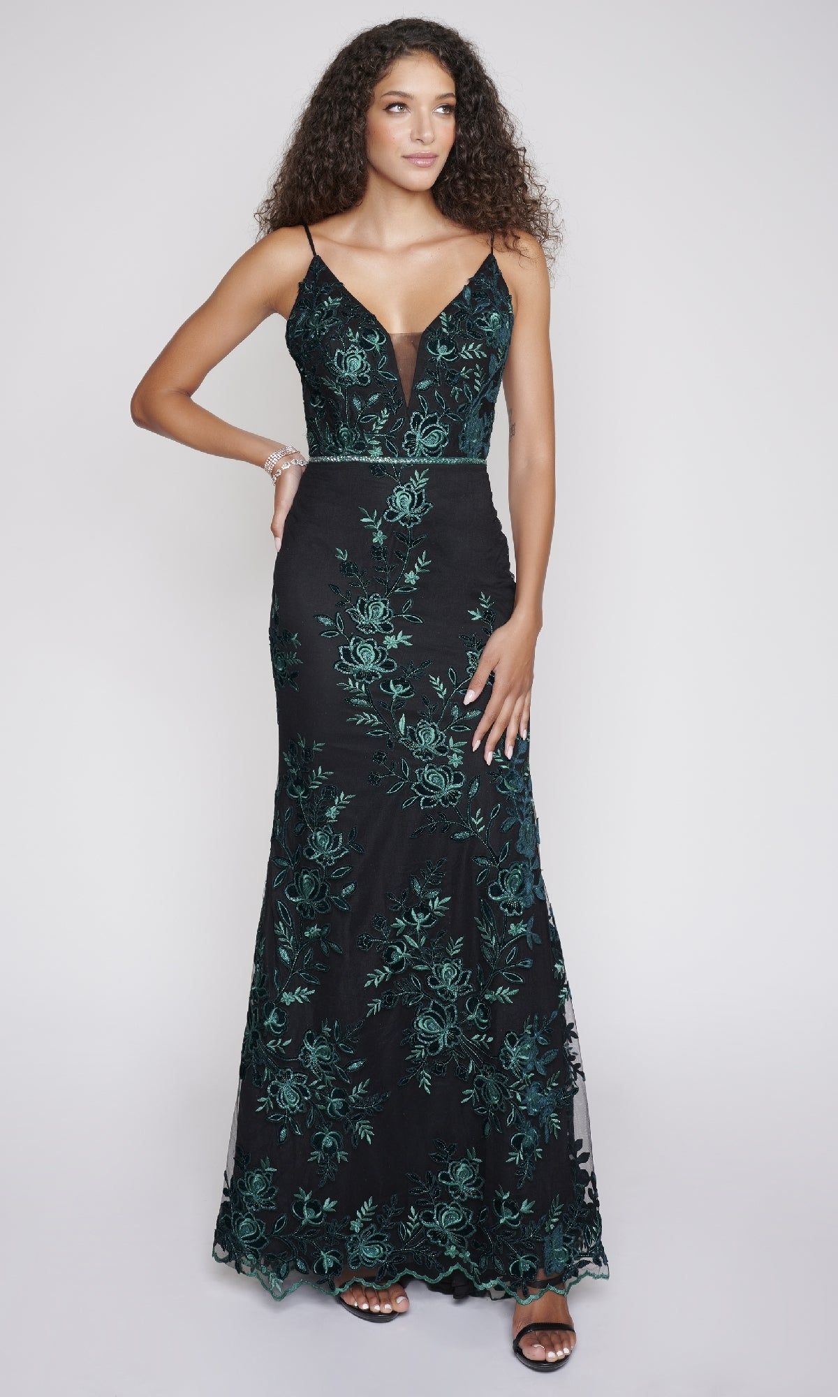 A woman models the Nina Canacci 2240 long formal dress—a sleeveless black gown with green floral embroidery, deep V neckline, mermaid silhouette—and pairs it with black high-heeled sandals against a plain background.