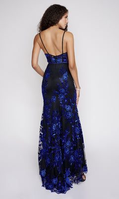 A woman models the Long Formal Dress: Nina Canacci 2240, showcasing its mermaid silhouette, blue floral patterns on black fabric, open back, and thin straps against a plain light background.