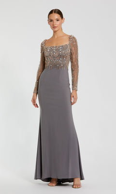 A woman models the Mac Duggal 2256 Long Sleeve Long Formal Dress, a grey floor-length gown with a fitted bodice, sheer sleeves, and metallic embellishments, standing against a plain background.