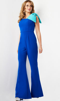 A woman stands against a plain white background, dressed in the Long Jumpsuit: Jovani 22589, an elegant royal blue jumpsuit with wide legs and a striking large light blue bow accent on one shoulder.