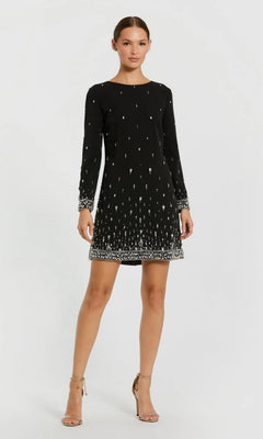 A woman stands with one hand on her hip and a neutral expression, wearing the Mac Duggal 2262 Short Semi-Formal Dress—a black, long-sleeve mini dress with crystal beading.
