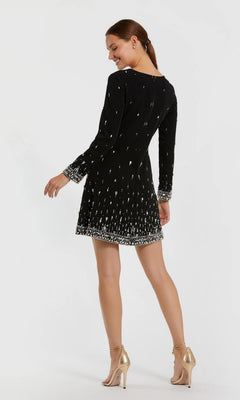 A woman in the Mac Duggal 2262 short semi-formal dress with long sleeves, black fabric, white patterns, and metallic high-heeled sandals stands facing away with her head turned, set against a plain white background.