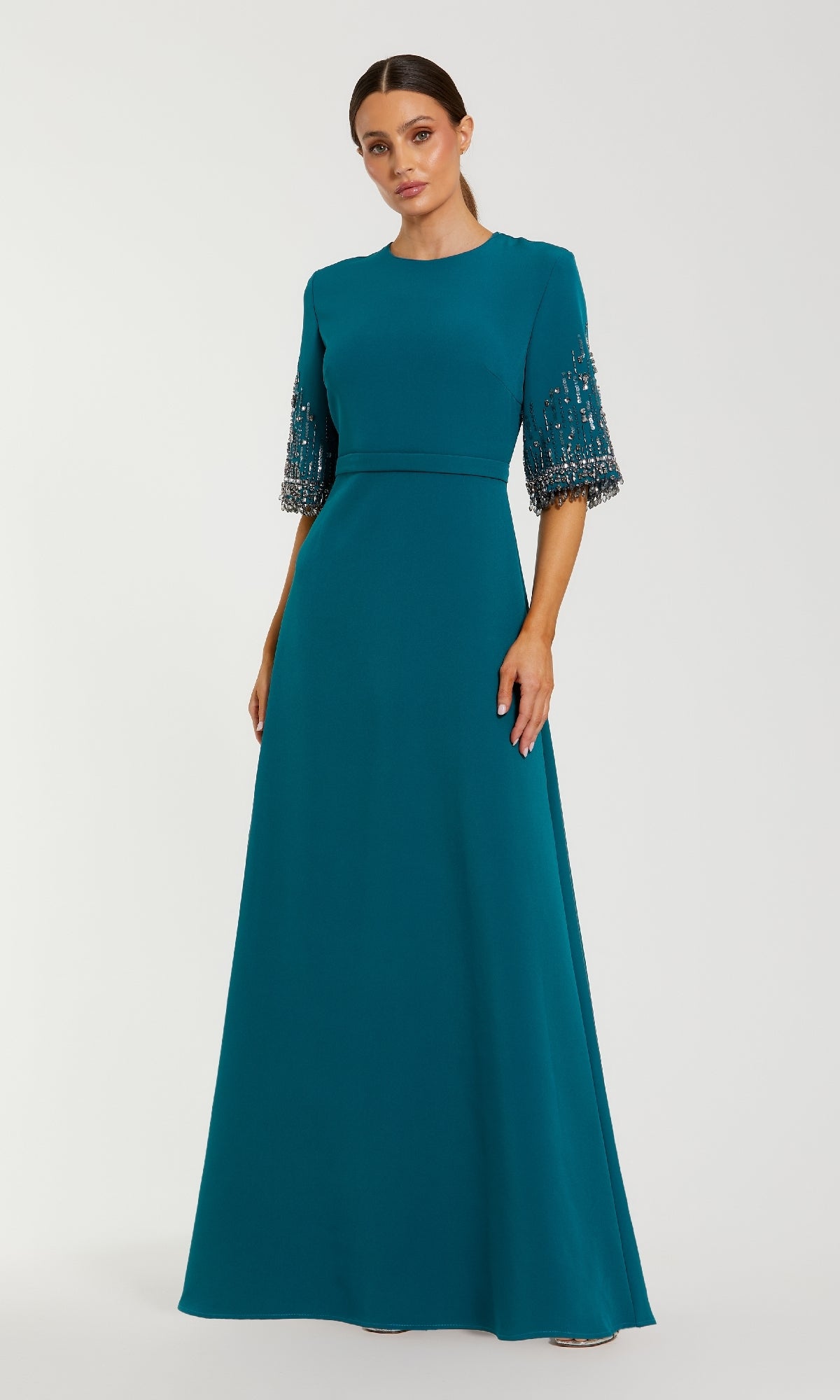 A woman wears the Mac Duggal 2270 Short Sleeve Sleek Formal Dress, a floor-length teal gown with bead and sequin accents, standing against a plain light background.