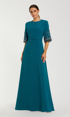 A woman wears the Mac Duggal 2270 Short Sleeve Sleek Formal Dress, a floor-length teal gown with bead and sequin accents, standing against a plain light background.
