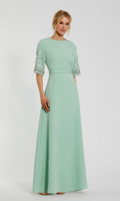 A woman in a light green Mac Duggal 2270 Short Sleeve Sleek Formal Dress, featuring sparkling bead embellishments, stands against a white background.