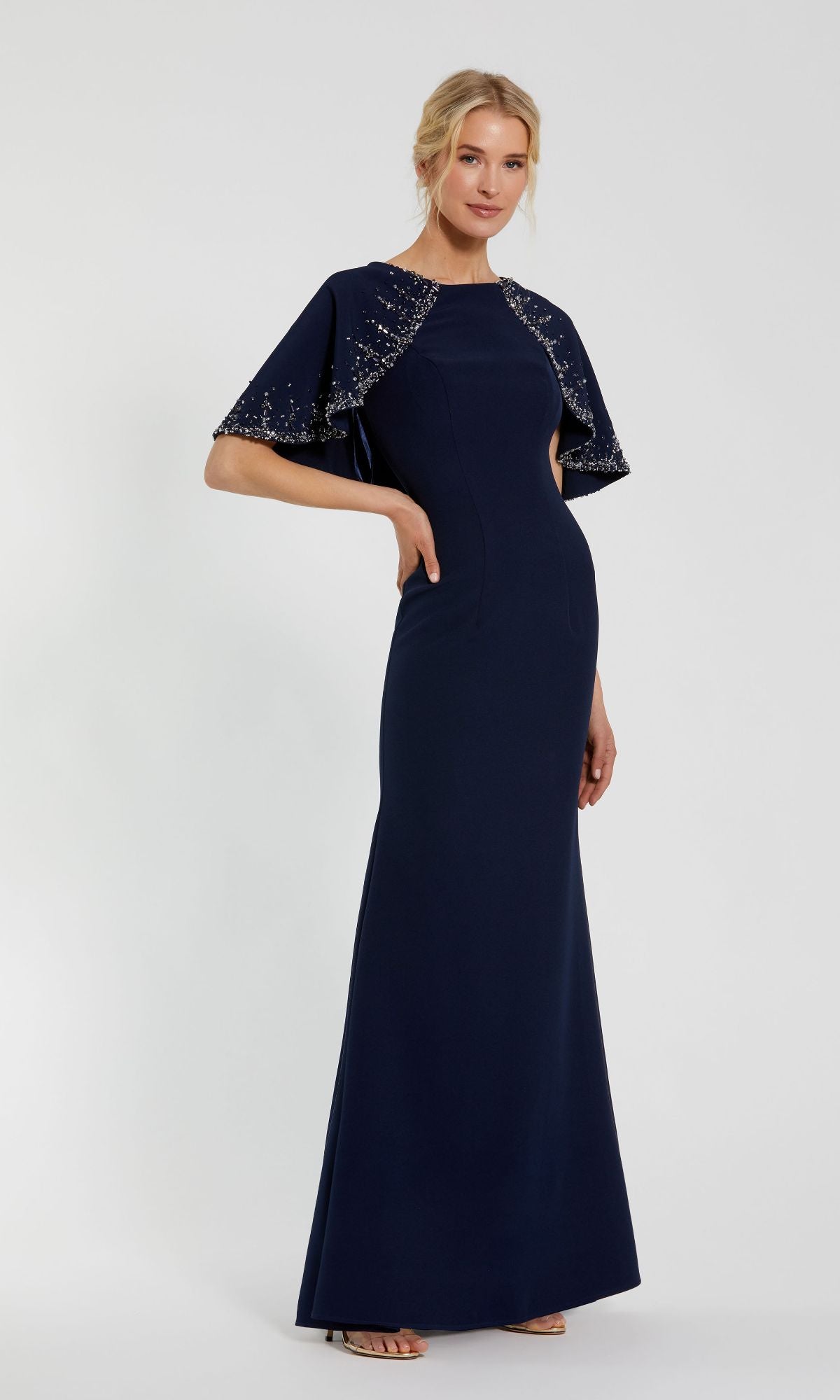 A woman poses in the Mac Duggal 2271 Caped Long Blue Formal Dress—floor-length, fitted, with beaded cape sleeves—an elegant mother of the bride choice, set against a plain light background.