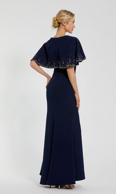 A woman models the Mac Duggal 2271 Caped Long Blue Formal Dress with a beaded capelet, looking to the side—an elegant mother of the bride option shown against a plain background.