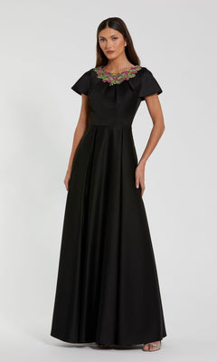 A woman poses against a plain white background in the Mac Duggal 2272 short sleeve black formal dress, featuring a modest floor-length design and a colorful beaded neckline.