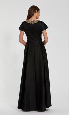 A woman stands with her back to the camera, wearing the Mac Duggal 2272 Short Sleeve Black Formal Dress, a floor-length gown with short sleeves and vibrant floral embroidery on the upper back.