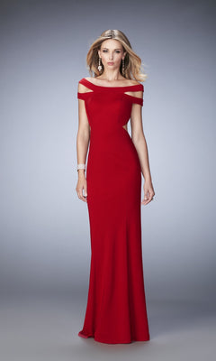 A woman poses on a gray background wearing the La Femme Dress 22728, a floor-length, off-the-shoulder gown featuring cut-out shoulder details and a strappy neckline, accessorized with earrings and a bracelet.