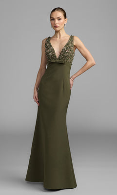 A woman stands against a plain gray background wearing the Mac Duggal 2278 Beaded-Bodice Long Prom Dress in olive, featuring a floor-length silhouette and deep V-neckline adorned with sparkling beadwork.