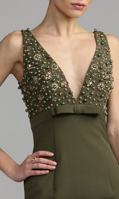 A woman wearing the Mac Duggal 2278 Beaded-Bodice Long Prom Dress in olive green, featuring a sleeveless empire waist, deep V-neckline, pearl and gem embellished bodice, a bow accent at the waist, and a ring on her right hand.