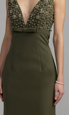 A woman wearing the Mac Duggal 2278 Beaded-Bodice Long Prom Dress—an olive green, sleeveless evening gown featuring a deep V-neckline, intricately beaded bodice, and a small bow accent at the waist—paired with a pearl bracelet on her right wrist.