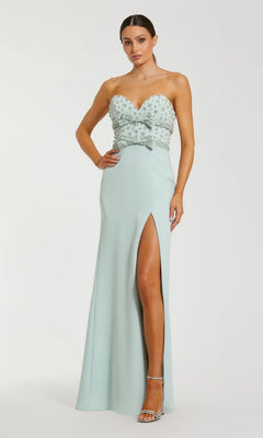 A woman stands against a plain light background in the Mac Duggal 2285 Strapless Sweetheart Formal Gown in mint, featuring a beaded bodice, front bow detail, high slit, and silver heels.