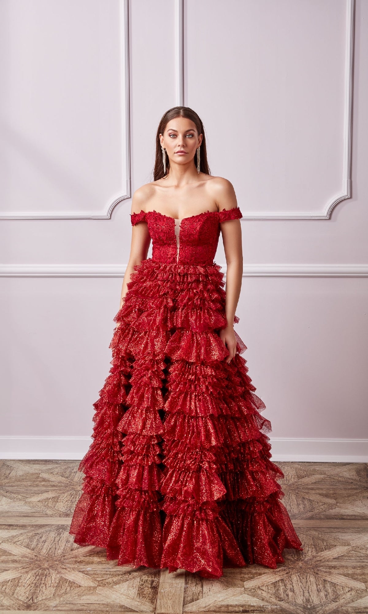 A woman stands indoors against a light pink paneled wall and wooden floor, wearing a Lace-Up Long A-Line Glitter Prom Dress with Ruffles.