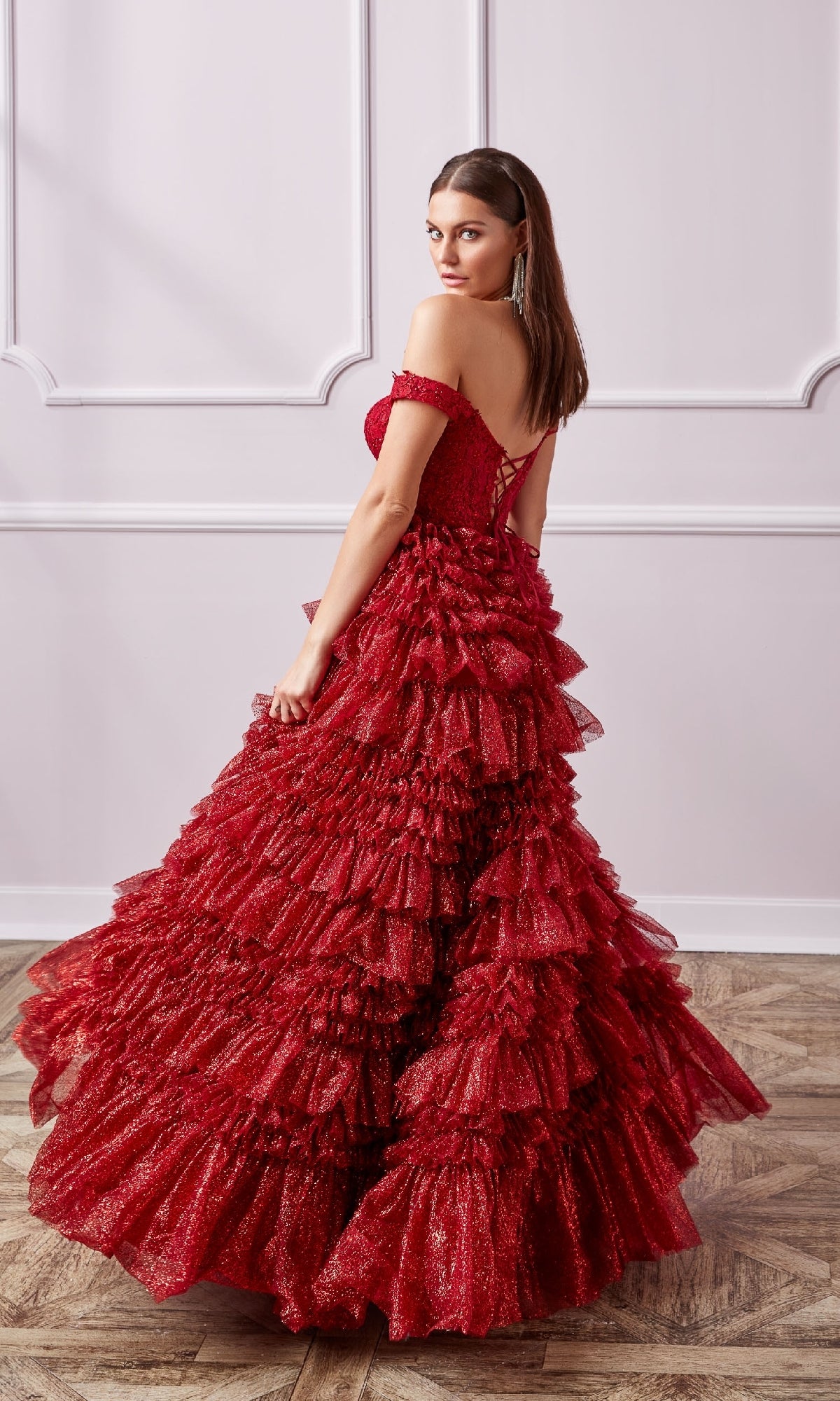 A woman stands on a wooden floor wearing the Lace-Up Long A-Line Glitter Prom Dress with Ruffles in red, featuring an off-the-shoulder neckline and sparkling glitter details, as she looks over her shoulder.