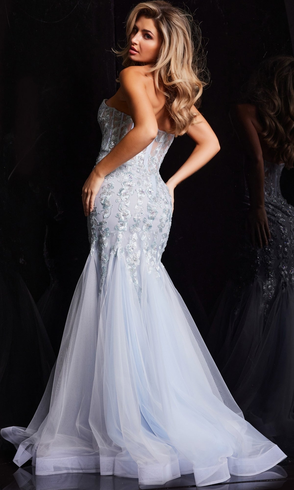 A woman poses in the Jovani Strapless Long Light Blue Prom Dress 22924, featuring floral embellishments and a tulle mermaid train, set against a dark background.
