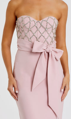 A woman wears the Mac Duggal 2294 Strapless Sweetheart Formal Gown in light pink, featuring a beaded geometric bodice and large waist bow—an elegant look for any formal event.