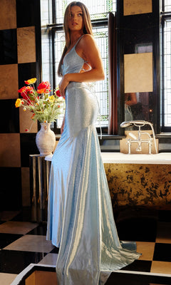A woman in the Jovani Strappy-Back Long Beaded Prom Dress 23010 in light blue stands by a bathtub in a bathroom with black and beige checkered tiles; a vase of red and yellow flowers rests on a nearby table.