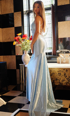 A woman in a light blue Jovani Strappy-Back Long Beaded Prom Dress 23010 stands in a tiled bathroom near a bathtub, with a vase of red and yellow flowers on the counter beside her.