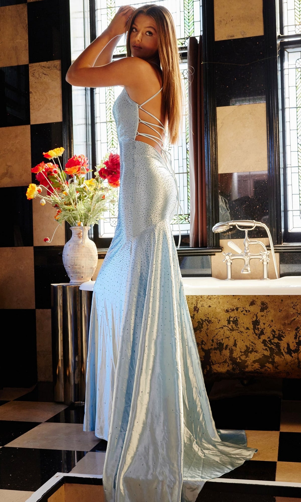 A woman wearing the Jovani Strappy-Back Long Beaded Prom Dress 23010 stands by a bathtub with a vase of flowers, in a room featuring checkered floors and a stained-glass window.