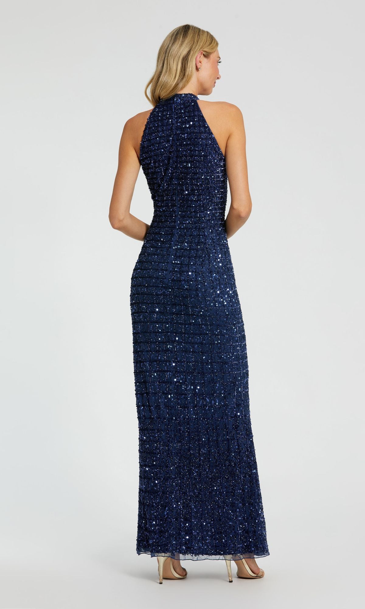 A woman faces away, wearing the Mac Duggal 23018 high-neck long beaded prom dress in navy blue with a halter neckline and sleeveless design, paired with beige high heels, set against a plain background.