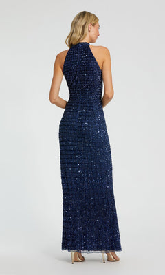 A woman faces away, wearing the Mac Duggal 23018 high-neck long beaded prom dress in navy blue with a halter neckline and sleeveless design, paired with beige high heels, set against a plain background.