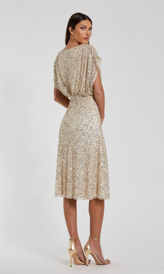 A woman is standing in the Mac Duggal 23027 knee length party dress, featuring beige sequins, an elegant a-line skirt, and short sleeves. She pairs it with gold high-heeled sandals and faces slightly away from the camera against a plain background.