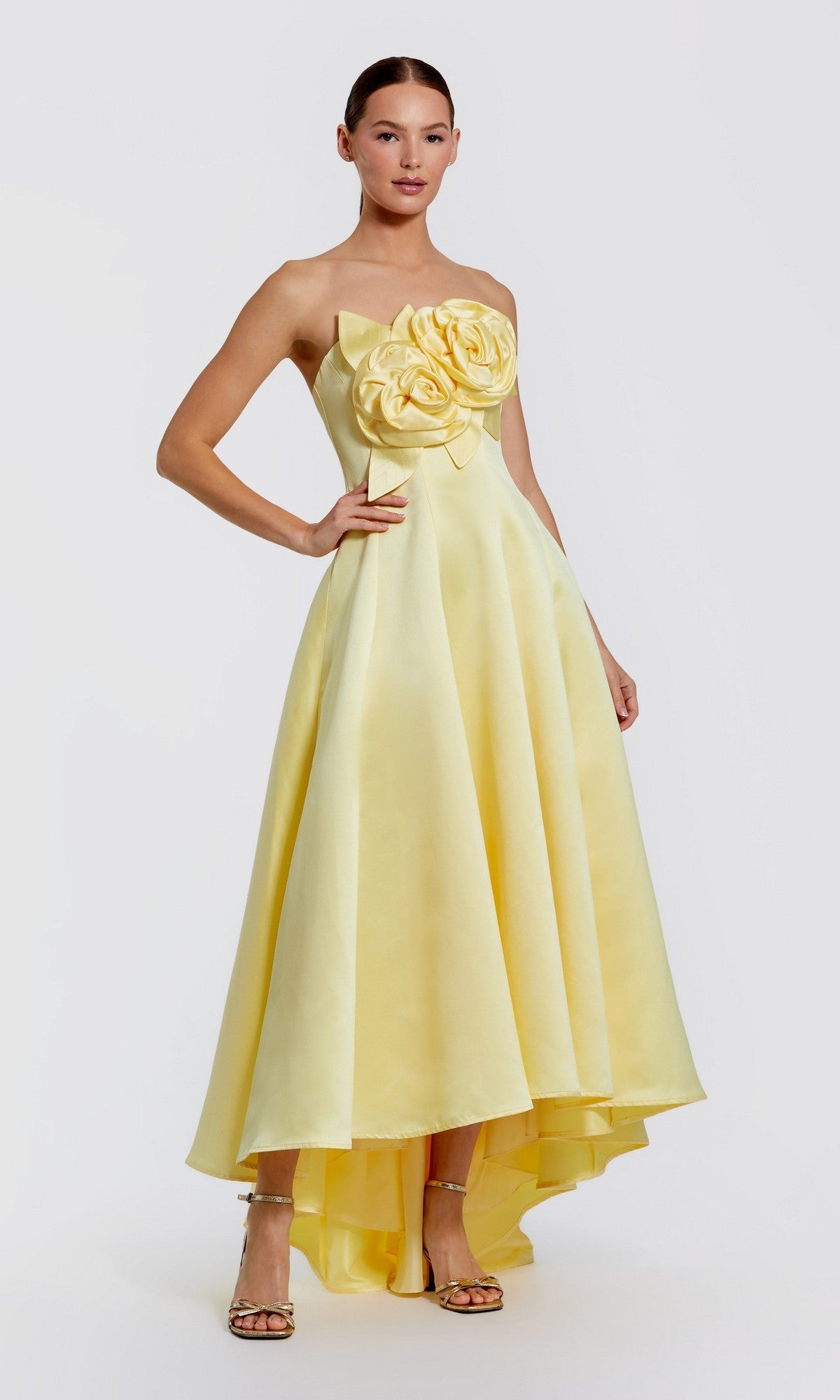 A woman models the Mac Duggal 2303 High-Low Formal Dress in Buttercup, featuring a strapless satin bodice with large 3D flower embellishments and a high-low hemline, styled with strappy heels against a plain light background.
