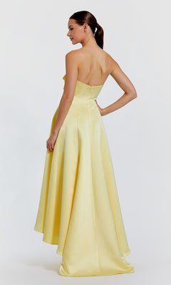 A woman models the Mac Duggal 2303 High-Low Formal Dress in yellow satin, strapless and floor-length, standing with her back to the camera and head turned to the side against a plain white background.