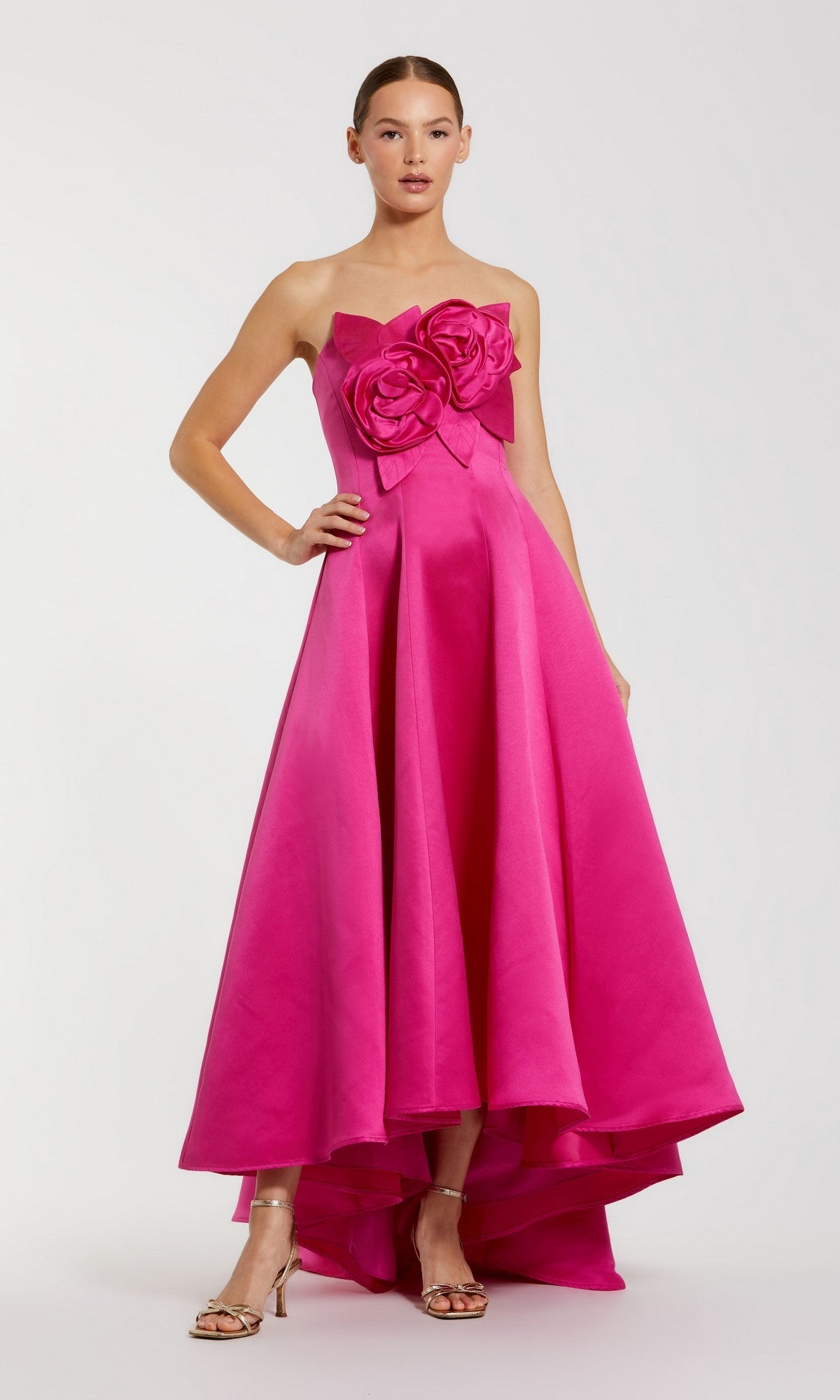 A woman in the Mac Duggal 2303 High-Low Formal Dress in hot pink, featuring a strapless satin bodice with bold 3D flower accents and a high-low hem, pairs it with metallic heels against a plain light background.